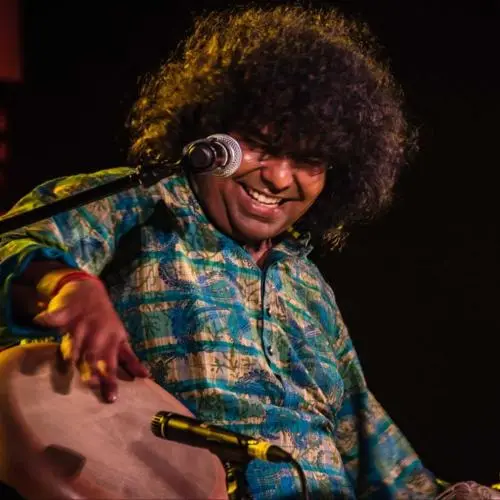Percussionist performing on stage, smiling while playing a hand drum, with a microphone and cymbals visible under warm stage lighting.