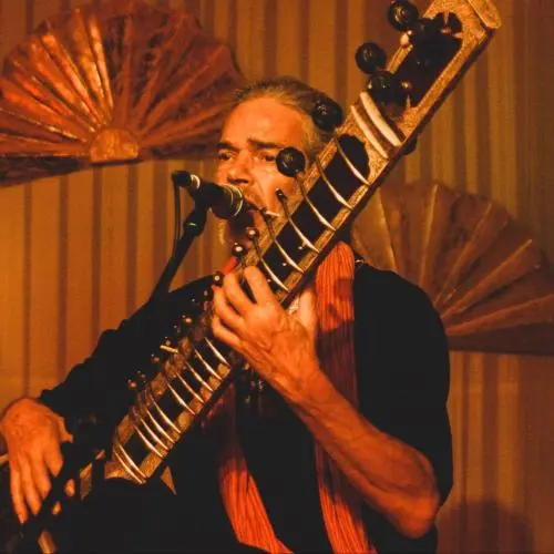 Color photograph of a male musician performing on stage, singing into a microphone while playing a sitar. He is wearing a dark outfit with a scarf, and the scene is lit with warm, amber lighting. Decorative fan-shaped elements are visible in the background, enhancing the stage setting.