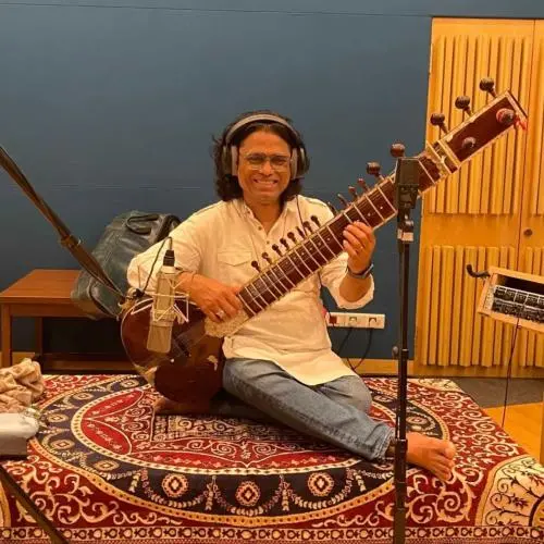 Sitar player Ravi Chary seated in a recording studio, playing the instrument with microphones set up around him and wearing headphones.