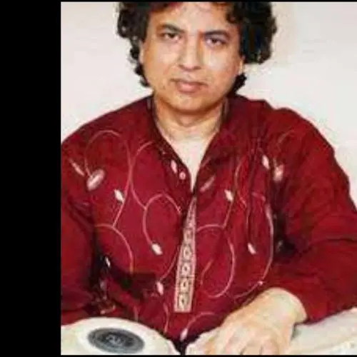 Color photograph of a male musician seated with a tabla, looking directly at the camera with a composed expression. He is wearing a red patterned kurta, and the image focuses on his upper body and the instrument, suggesting a posed portrait rather than a live performance setting.