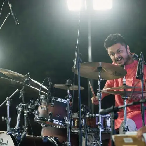 Percussionist in a pink kurta performing on a drum kit on stage, surrounded by microphones and stage lights during a live music performance.