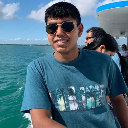 Young man wearing sunglasses and a blue T-shirt standing on a boat, with ocean water and a partly cloudy sky in the background.