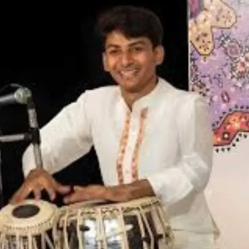 A tabla player in a white kurta performing on stage, smiling while playing the tabla with a microphone placed nearby.