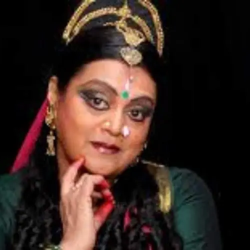 A close-up portrait of classical dancer Swapnasundari in traditional Kuchipudi/Bharatanatyam attire, featuring ornate gold headgear, heavy eye makeup, and red-stained fingers held to her face against a black background.