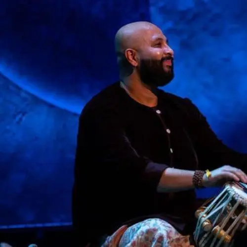 A tabla player performing on stage under dramatic blue lighting, seated behind a pair of tablas during a live Indian classical music concert.