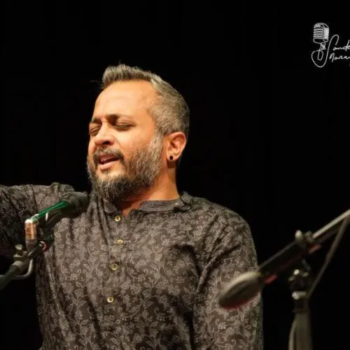 Carnatic vocalist performing on stage, singing with raised hand gesture and microphone.