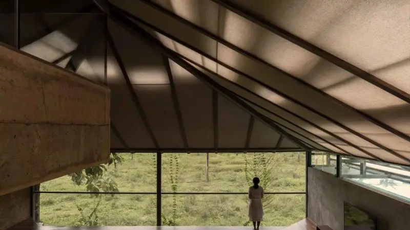 A spacious modern interior with sloped ceiling and large glass window, overlooking greenery with a person standing near the edge.