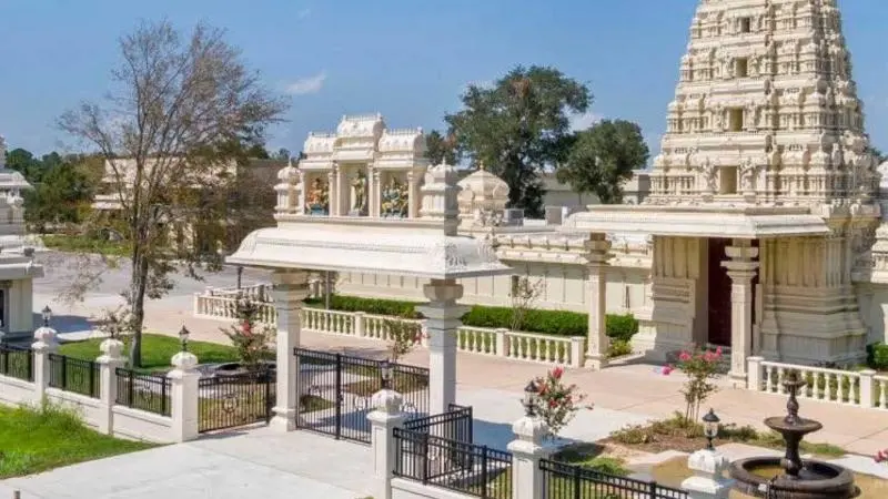 Exterior view of Meenakshi Temple complex in Houston featuring ornate white gopuram tower, temple structures, entrance gate, and landscaped surroundings under a clear sky.
