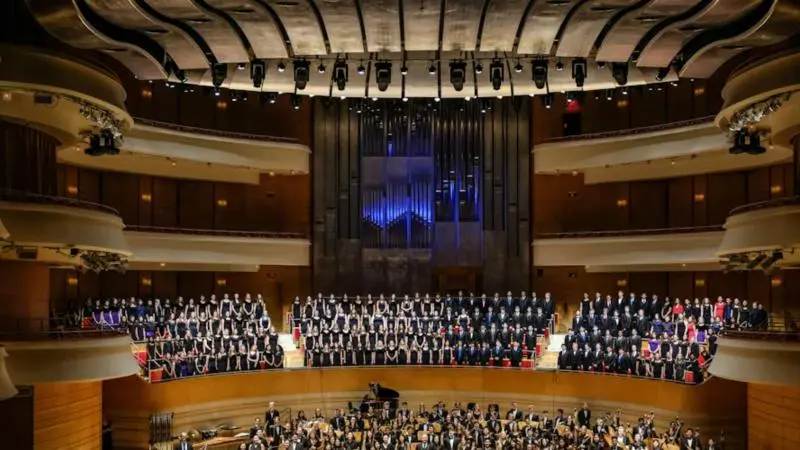 Interior view of Capistrano Concert Hall featuring a full orchestra and large choir assembled on stage, with tiered balcony seating, warm wooden interiors, and a prominent pipe organ at the back under modern acoustic ceiling panels.