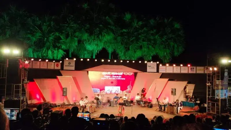 Open-air auditorium hosting a live music concert at night, with performers on a brightly lit stage, surrounded by palm trees and a large seated audience watching the performance.