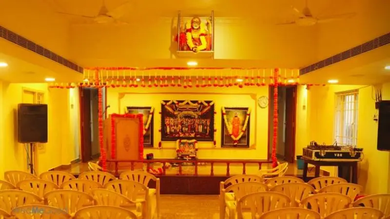 Interior view of Sri Krishna Saras Hall featuring a small decorated stage, devotional portraits, and rows of seating arranged for a cultural or spiritual gathering.